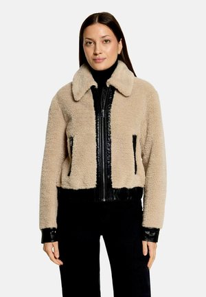 Vrouw draagt een beige sherpa-jas met zwarte leren rits- en zakdetails over een zwarte coltrui en zwarte broek.