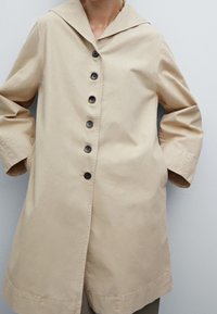 Manteau en coton beige avec un grand col, fermeture à boutons sur le devant et deux poches latérales. Il présente des manches longues et une coupe décontractée arrivant aux genoux.