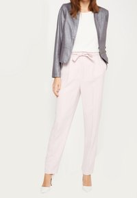 Blazer gris métallique sur un haut blanc, associé à un pantalon taille haute rose clair et des talons blancs, avec un détail de nœud noué.