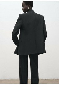 Traje negro a medida que presenta una blazer larga con solapas afiladas, hombros estructurados y pantalones de pierna ancha. Tejido suave con textura sutil.