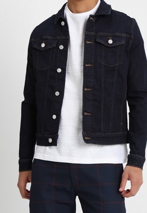 Mann trägt eine dunkelblaue Jeansjacke über einem weißen, strukturierten Hemd und dunkelblauen Hosen mit roten Karomustern.