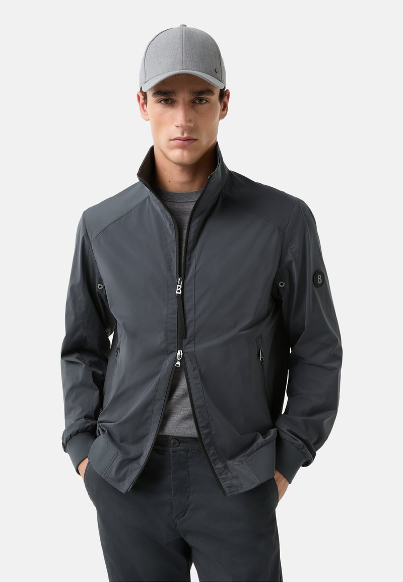 Dunkelgraue Zip-Jacke mit hohem Kragen, elastischen Bündchen und Seitentaschen. Getragen über einem grauen Hemd, kombiniert mit einer hellgrauen Mütze.