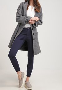 Lång grå ullkappa med svarta knappar, vit topp och marinblå smala byxor, tillsammans med ljusgrå sneakers.