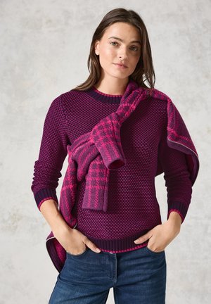 Pull en tricot violet avec un motif texturé, col rond et poignets côtelés, accompagné d'une écharpe à carreaux rose portée sur une épaule.