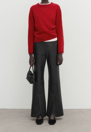 Personne portant un pull rouge vif, un pantalon noir à jambes larges avec des fentes sur le devant, des chaussures noires, tenant un petit sac à main noir tissé.