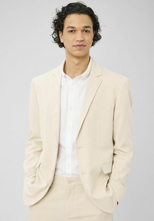 Jeune homme aux cheveux noirs bouclés portant un costume beige et une chemise blanche, mains dans les poches, fond gris neutre.