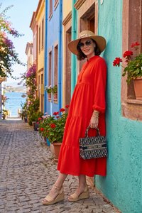 Mujer con vestido rojo y sombrero de sol apoyada en una pared turquesa en una calle empedrada con edificios coloridos y flores en macetas cerca del paseo marítimo.