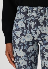 Jeans florali cu o bază de culoare bleumarin închis, decorate cu modele de trandafiri albaștri deschiși. Materialul pare texturat, având un design clasic cu cinci buzunare.