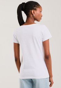 Žena s copánkovým culíkem, oblečená v jednoduchém bílém tričku s krátkým rukávem a světlých modrých džínách, otočená zády, na neutrálním pozadí.