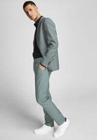 Completo grigio chiaro con una vestibilità slim, caratterizzato da un blazer a un bottone, abbinato a una camicia nera e sneakers bianche. Tessuto dalla texture liscia.