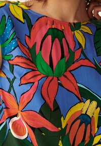 Un tessuto a motivo floreale presenta grandi fiori colorati in rosa, rosso e arancione su uno sfondo blu, con foglie verdi e dettagli.