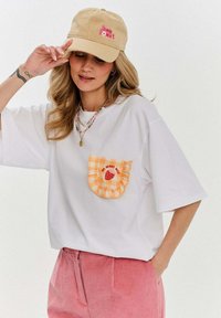 Bílé oversized tričko s žlutou kostkovanou kapsou, na které je design červené jahody. V kombinaci s růžovými manšestrovými kalhotami a béžovou kšiltovkou.