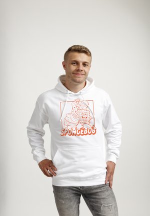 Junger Mann lächelnd, trägt weißen Hoodie mit orangefarbenen SpongeBob-, Patrick- und Gary-Figuren und grauen verwaschenen Jeans, steht entspannt.