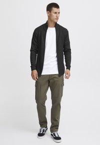 Jachetă neagră cu nervuri, cu fermoar, purtată peste un tricou alb, asortată cu pantaloni cargo verzi măslinii și adidași negri cu tălpi albe.