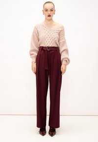 Maglione rosa lavorato a maglia con motivi a treccia, abbinato a pantaloni bordeaux a vita alta e scarpe bordeaux a punta con dettagli in metallo.