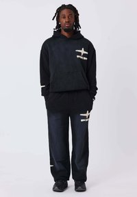 Sudadera negra con pantalones a juego, adornados con parches abstractos blancos. El material parece suave; la sudadera cuenta con un bolsillo frontal, y los pantalones tienen una cintura elástica.