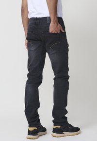 Vaqueros de mezclilla negra con un corte slim, que cuentan con bolsillos traseros y detalles de costura amarilla. Llevados con zapatillas negras.