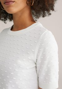 Närbild av en person som bär en vit, strukturerad kortärmad tröja med små upphöjda prickmönster och ringörhängen.