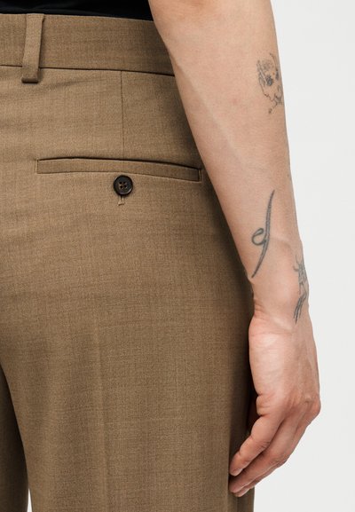 Κοντινό πλάνο του γοφού ενός ατόμου που δείχνει καφέ ραμμένα παντελόνια με κουμπωτή πίσω τσέπη και ένα τατουάζ στο αντιβράχιο που ακουμπά δίπλα.