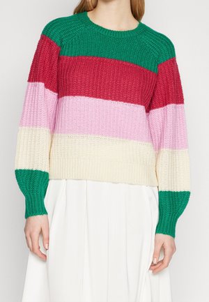 Frau trägt einen grob gestrickten Pullover mit horizontalen Streifen in Grün, Rot, Pink, Creme und Grün, kombiniert mit einem weißen Rock.