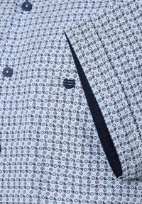 Koszula w wzory z geometrycznym wzorem w niebiesko-białe, z kieszonką i granatowymi akcentami. Guziki są ciemnoniebieskie. Gładka tekstura materiału.