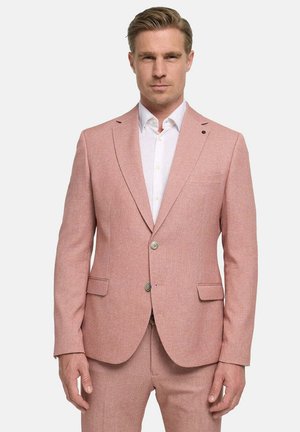 Hombre vestido con chaqueta de traje rosa a medida y pantalones a juego con camisa blanca, de pie frente a un fondo liso.