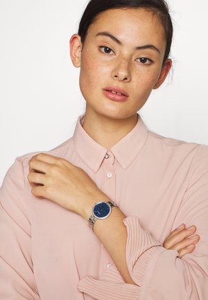 Silver wristwatch with a round blue speckled face, embellished bezel, and sleek band, worn on a hand over a light pink button-up shirt.