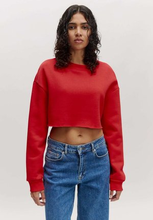 Roter cropped Sweatshirt mit langen Ärmeln, weicher Textur, geripptem Rundhalsausschnitt, kombiniert mit hoch taillierten blauen Jeans aus Denim, die vordere Taschen haben.