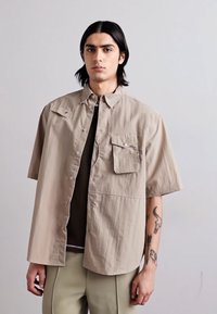Beige kortärmad skjorta med knäppning fram, stor bröstficka och avslappnad passform, bärs över en mörk topp. Slät tyg med minimal textur.