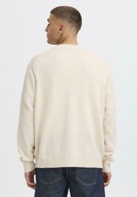 En mann med kort hår står med ryggen til, iført en lys beige genser og mørke jeans, mot en ensfarget hvit bakgrunn.