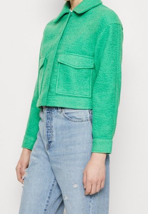 Personne portant une veste courte vert vif avec de grandes poches avant et un jean bleu clair taille haute déchiré.