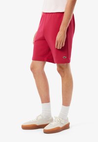 Pantalones cortos deportivos rojos hechos de un tejido suave, con una cinturilla elástica y un pequeño logo verde. Llevados con calcetines blancos y zapatos claros.
