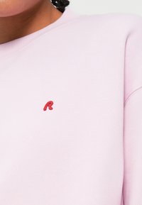 Ανοιχτό ροζ φούτερ φτιαγμένο από μαλακό ύφασμα, με πλεκτό λαιμό και ένα μικρό κοκκινωπό κεντημένο γράμμα 'R' στο στήθος.