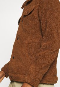 Chaqueta de forro polar marrón con superficie texturizada, que cuenta con un frontal abotonado, bolsillo en el pecho y bolsillos laterales. Ribeteada en los puños y el dobladillo.