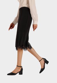 Scarpe con tacchi alti neri, con punta affusolata e cinturino alla caviglia. Finitura in pelle liscia, con tacco a blocco e design elegante. Dettaglio in pizzo sull'orlo della gonna.