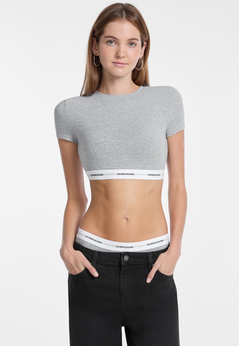 T-shirt gris court à manches courtes, présentant un design ajusté et une bande blanche avec "GUESS JEANS" imprimé en noir.