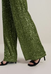 Pantalons larges en sequins verts avec une surface texturée, assortis à des talons ouverts noirs, mettant en valeur une taille ajustée et une silhouette ample.