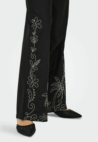 Črne široke hlače z belim cvetličnim vezenjem ob straneh. Kombinirane z črnimi tkanimi muleci s koničastimi prsti.