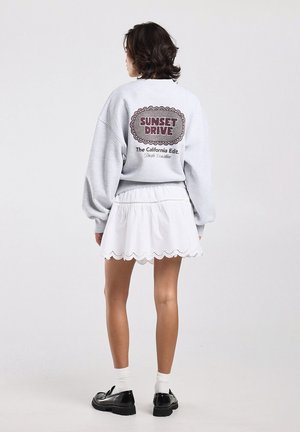 Femme aux cheveux bouclés courts portant un sweat-shirt gris avec le texte « Sunset Drive », une jupe blanche à bord festonné, des chaussures noires et des chaussettes blanches.
