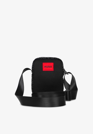 Sac bandoulière en mesh noir avec une sangle réglable et un patch rouge marqué HUGO à l'avant, isolé sur fond blanc.