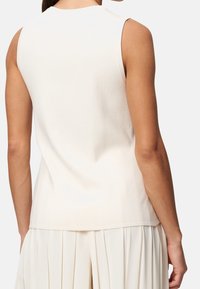 Femme portant un haut blanc sans manches et une jupe plissée blanche, vue de dos contre un fond blanc uni.