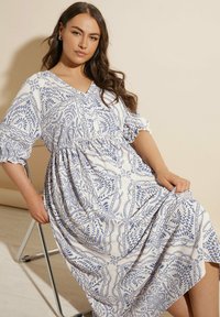 Valkoinen maksimekko, jossa on sininen kukkakuosi, v-pääkolo ja lyhyet puffihihat. Kankaan pinta on teksturoitu, vyötärö on kasattu ja helma laskeutuu elävästi.