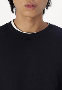 Navask blå sweatshirt med ribbet rund hals, blød tekstur og en lagdelt hvid skjorte synlig ved kraven. Ingen grafik eller mønstre.