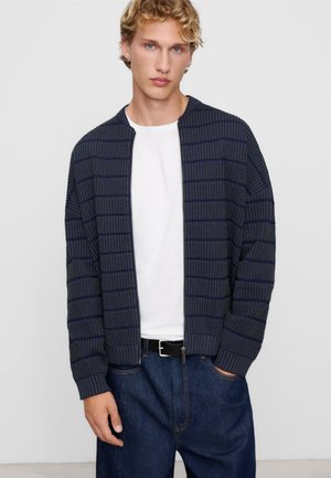 Młody mężczyzna z kręconymi blond włosami, ubrany w granatowy, kratkowany sweter na zamek błyskawiczny, na biały shirt i ciemnoniebieskie jeansy z czarnym paskiem.