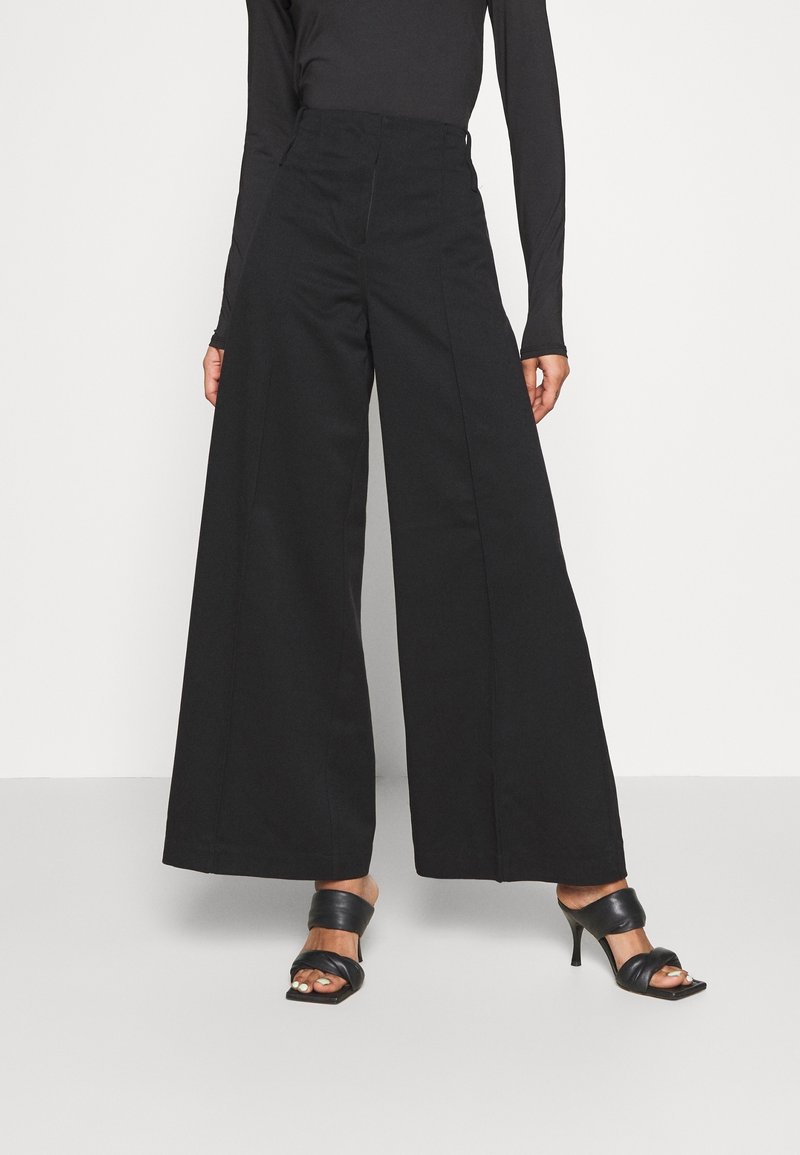 Czarne spodnie z szerokimi nogawkami i wysokim stanem, wykonane z solidnego materiału, z przodu plisowane i wykończone w minimalistyczny sposób. W zestawie z czarnymi sandałami na obcasie.