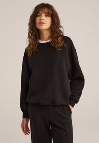 Felpa nera con scollatura a costine e maniche lunghe. Tessuto morbido, vestibilità rilassata, con polsini e fascia in vita, abbinata a pantaloni neri.