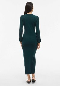 Vestido de punto ajustado en color teal, con mangas largas, escote redondo y corte recto hasta la mitad de la pantorrilla. Sin patrones visibles ni elementos adicionales.