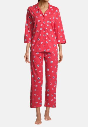 Person iført rød pyjamasæt med hvidt og blåt blomstermønster, knappet top med krave og bukser med lige ben, stående barfodet.