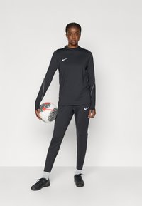 Μαύρη αθλητική μπλούζα με μακριά μανίκια και ταιριαστά παντελόνια με λευκές λεπτομέρειες. Μαύρα παπούτσια και μια μπάλα ποδοσφαίρου με σχέδιο σε μαύρο και άσπρο.