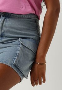 Denim mini-jurk met een lichtblauwe wassing, voorzien van zijzakken en een subtiele A-lijnvorm. Gecombineerd met een slanke zilveren armband en een roze ring.